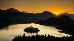 Sunrise view across Lake Bled to the island church and clifftop castle from Ojstrica as the run rises beside Storzic and Kocna mountains, Slovenia.