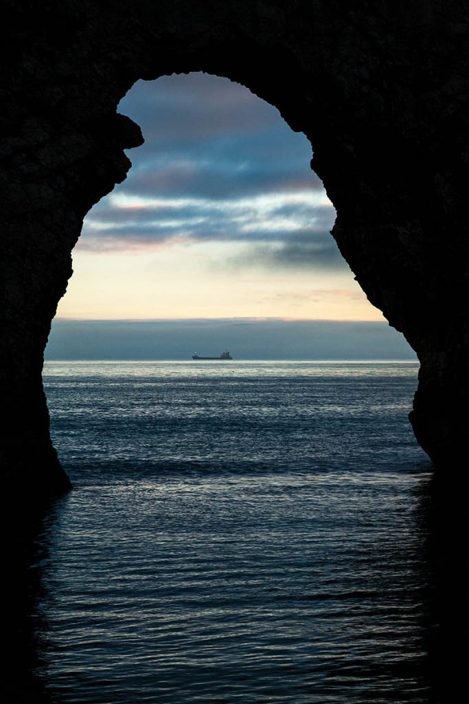 A view through the Durdle Door arch as a cargo ship sails by in the distance, Dorset, England. Durdle door is one of the many stunning locations to visit on the Jurassic coast in southern England.