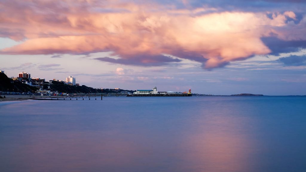 Bournemouth Pier and seafront as a storm cloud drifts over as is illuminated pink by the sunset. Highcliffe can also be seen on the far right.
