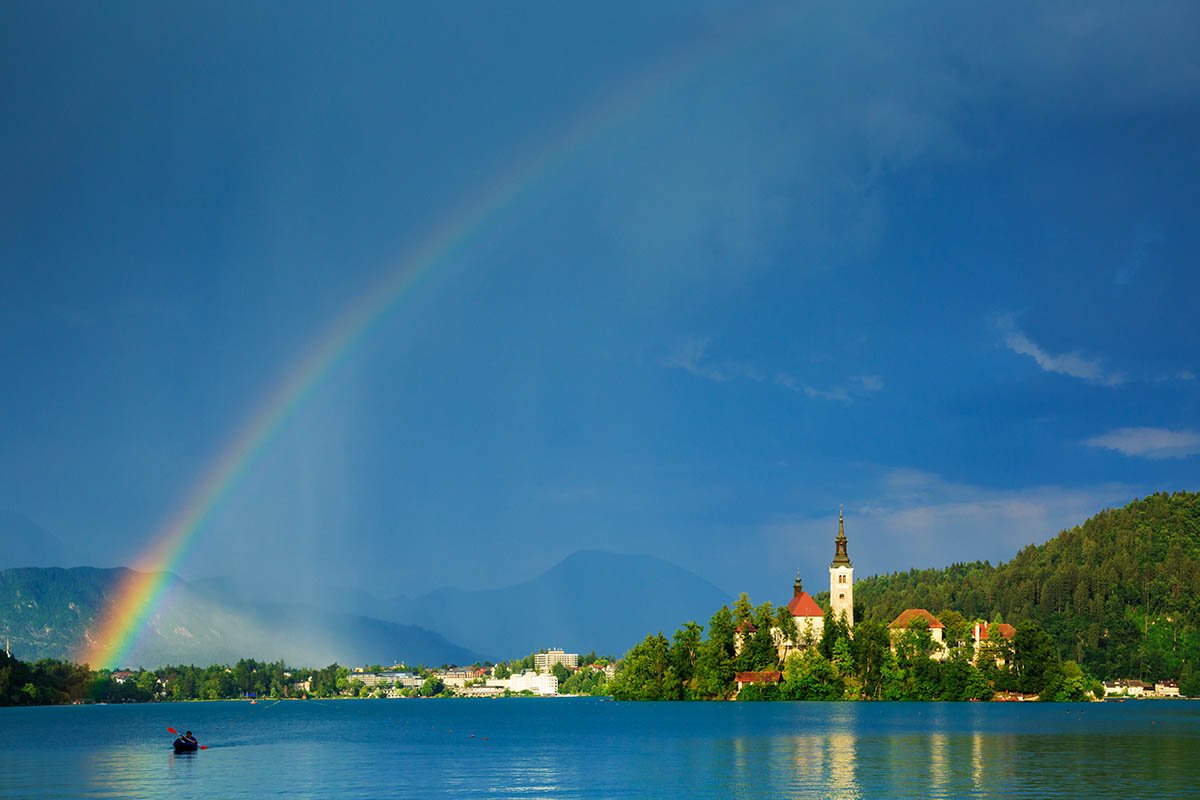 Beautiful light across to the beautiful Lake Bleds island church as a storm blows in and produces a rainbow right over the church and surrounding hills, Slovenia.