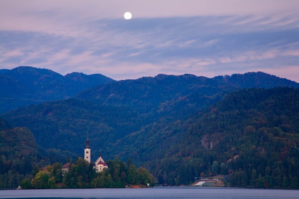 Moon setting at sunrise over hills and Island church at Lake Bled, Slovenia.