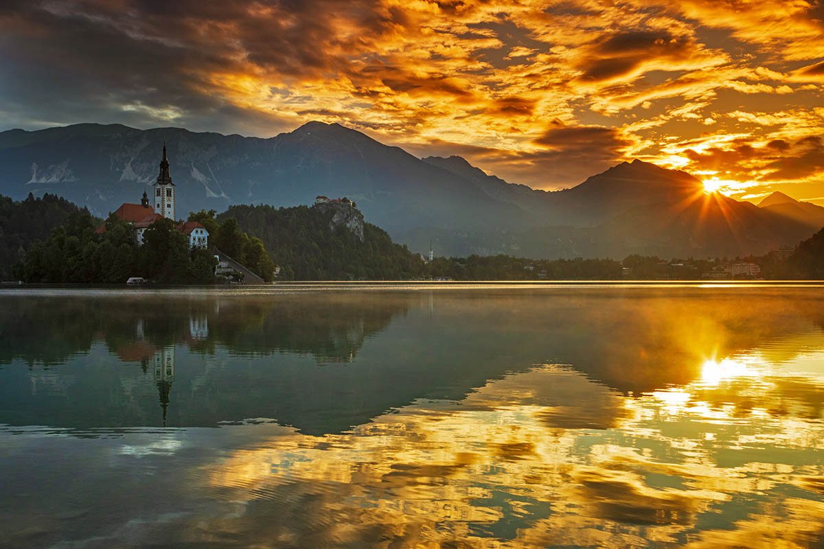 Summer sun rising over Karavanke mountains behind Lake Bled, island church of the assumption of Mary and castle, Slovenia.