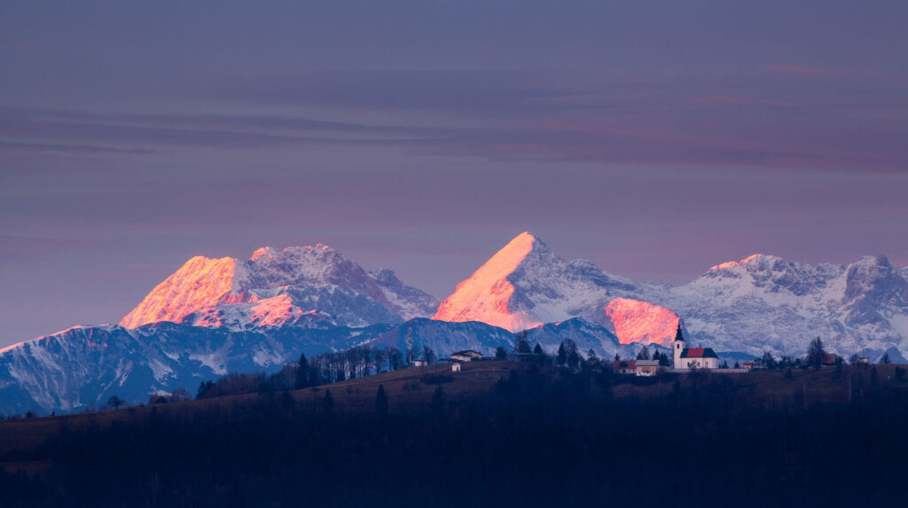 View across to the church of Saint Nicholas and the village of Jance at sunset with the Kamnik Alps in the background, in the hilly region to the east of Ljubljana, Slovenia. Seen from Mali Vrh village.