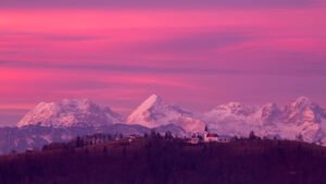 View across to the church of Saint Nicholas and the village of Jance at sunset with the Kamnik Alps in the background, in the hilly region to the east of Ljubljana, Slovenia. Seen from Mali Vrh village.